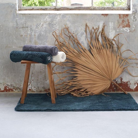 Trois tapis de bain roulés, gris foncé, bleus et blancs, sur un tabouret en bois.