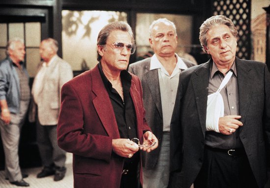 Drie mannen in pak staan bij elkaar. Eén man draagt een rode blazer, de ander een zwarte jas met verband om zijn arm.