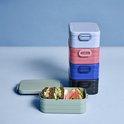 Pile de boîtes à lunch de couleurs variées : bleu clair, rose, bleu, noir et vert avec du pain et de la pastèque.