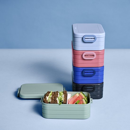 Pile de boîtes à lunch de couleurs variées : bleu clair, rose, bleu, noir et vert avec du pain et de la pastèque.