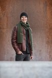 Jonge man met een bruine leren jas, zwart shirt, grijze jeans, zwarte muts en olijfgroene sjaal.