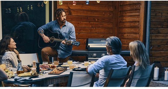 Mensen genieten van een maaltijd aan een tafel buiten, met een man die gitaar speelt en een barbecue op de achtergrond.