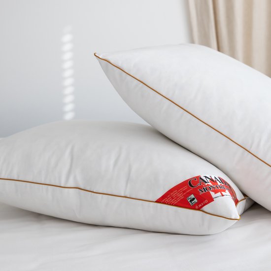 Twee witte kussens met bruine biezen op een bed. Een rode "Canada Montreal" label is zichtbaar op de onderste kussen.