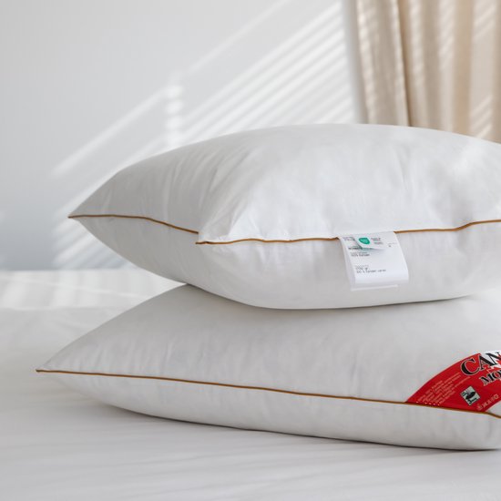 Twee witte kussens met bruine biezen op een bed. Een label is zichtbaar op de bovenste kussen.