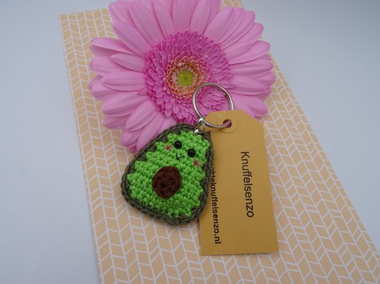 Gehaakte avocado sleutelhanger met gezichtje naast een roze bloem.