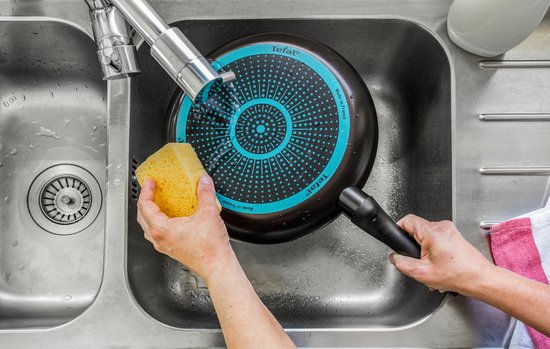 Een hand wast een pan onder de kraan met een spons. De onderkant van de pan heeft een blauw cirkelvormig antislipmotief.