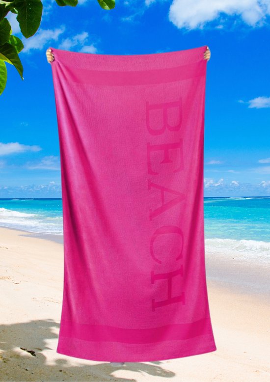 Een felroze strandlaken met de tekst 'BEACH' in reliëf wordt door twee handen vastgehouden tegen een blauwe lucht en zee.