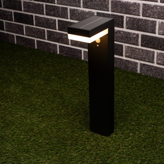 Zwarte solar tuinpaal met bewegingssensor op kunstgras naast een stenen muur.