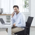 Man met een lichtblauwe blouse en beige broek zit op een stoel en belt met zijn telefoon.