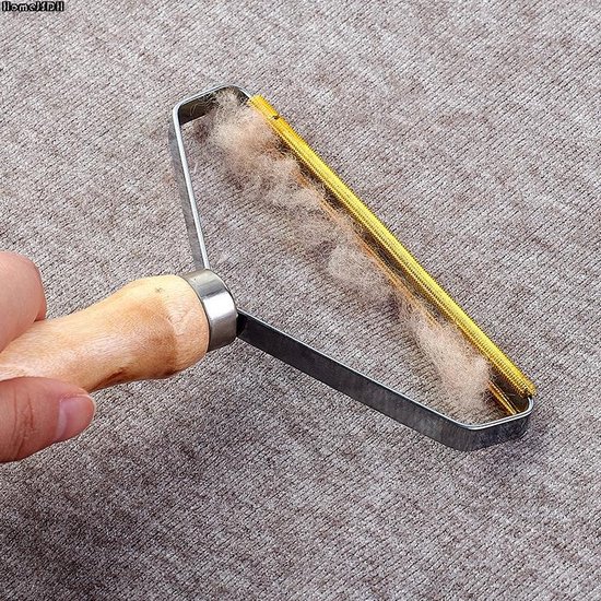 Hand gebruikt een pluizenroller op een grijze stof. Het apparaat is een pluizenverwijderaar met een metalen frame, gouden spiraalvormige koppen en een houten handvat.