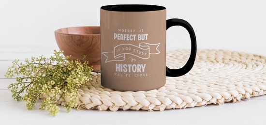 Tasse marron avec bordure et anse noires, portant le texte "NOBODY IS PERFECT BUT IF YOU STUDY HISTORY YOU'RE CLOSE", sur un plateau