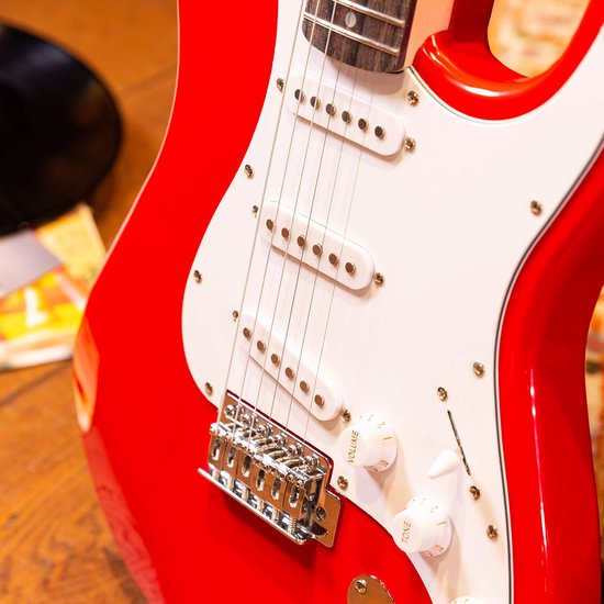 Detail van de rode elektrische gitaar, met witte slagplaat, pick-ups en knoppen.