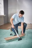 Man knielt op een groene ondergrond en gebruikt een laminaatsnijder om een houten plank door te snijden.