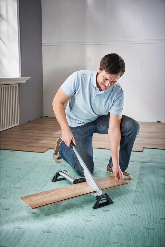 Man knielt op een groene ondergrond en gebruikt een laminaatsnijder om een houten plank door te snijden.