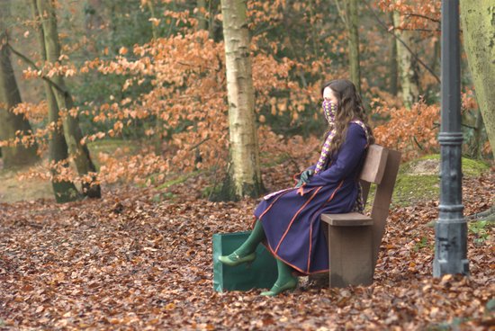 Christina Ricci met sjaal en jurk zit op een bankje in een herfstbos.