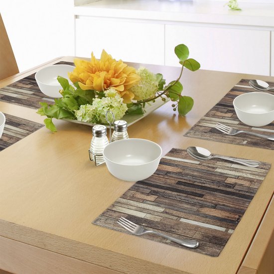 Table à manger avec des sets de table carrés à imprimé planches de bois, assiettes blanches, couverts et arrangement floral.
