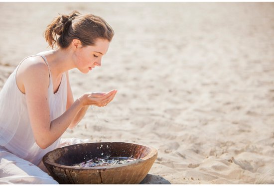 Vrouw met witte jurk op het strand, schept water op uit een houten kom.