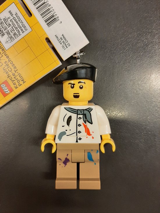 Lego-kunstenaarssleutelhanger met baret, wit schildershemd met verfvlekken en beige broek. Verpakking met productinformatie.