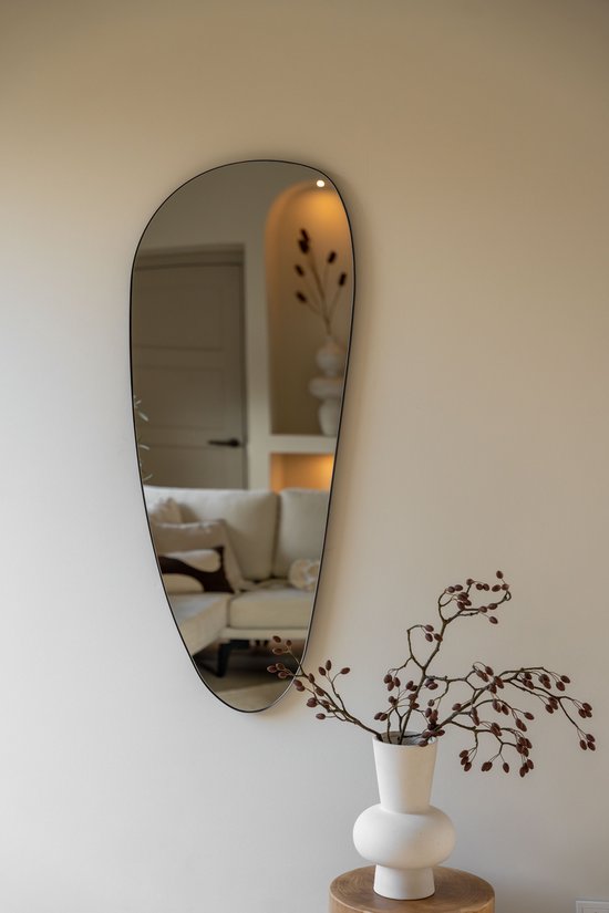 Organische zwarte spiegel met slanke lijst aan een lichte muur. De spiegel reflecteert een bank met kussens en een halfronde nis.