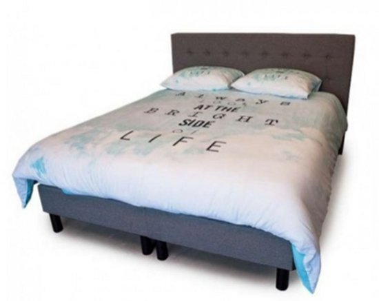 Grijze boxspring met gestoffeerd hoofdbord, blauw/wit dekbedovertrek met tekst 'ALWAYS AT THE BRIGHT SIDE OF LIFE' en kussens.