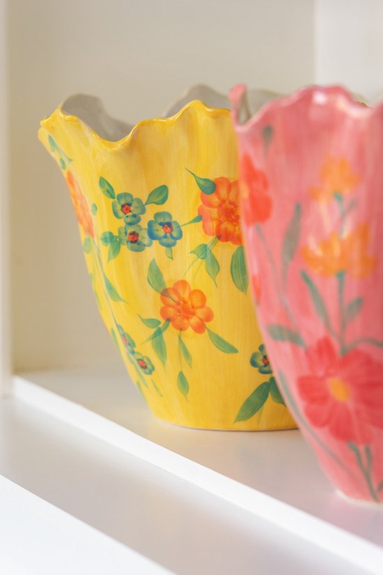 Vase jaune à bord ondulé, décoré de fleurs oranges et de petites feuilles vertes. Vase avec des fleurs roses à l'arrière-plan.