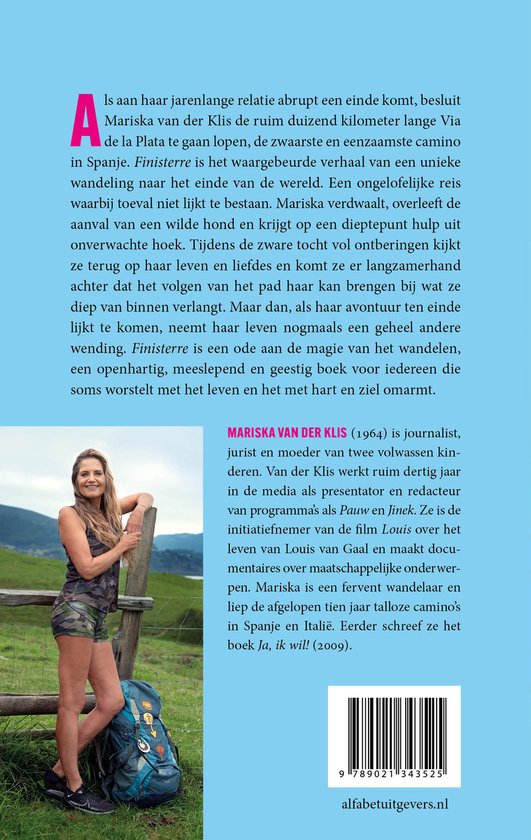 Foto van Mariska van der Klis, een journalist, presentator en redacteur, met camouflagekleding en een wandelrugzak, staand voor een houten hek in een bergachtig gebied.