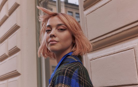 Model met oranje haar, draagt een geruite jas, kijkt naar de camera.