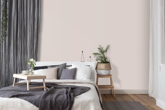 Een slaapkamer met een gestyled bed, lichte roze muren, grijze gordijnen en een houten bijzettafel met plant.