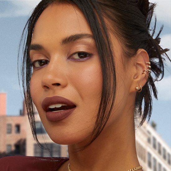 Model met donker haar en opgestoken kapsel, draagt donkerbruine lippenstift en meerdere oorbellen.