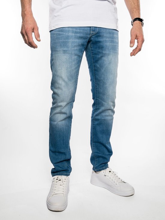 Lichtblauwe jeans met een slim fit pasvorm. Op de achtergrond een witte wand. De broek is voorzien van witte schoenen en een horloge.