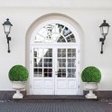 Dubbele witte deuren met glas-in-lood bovenlicht, geflankeerd door twee bolvormige buxusplanten in sierlijke potten.