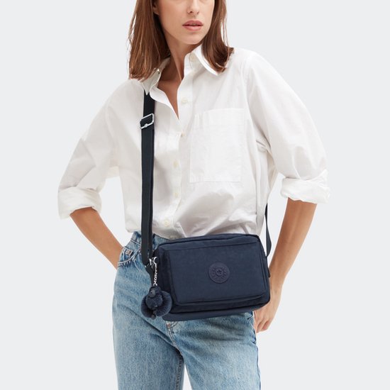Model met witte blouse en spijkerbroek draagt een donkerblauwe Kipling schoudertas met pompon-sleutelhanger.