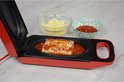 Rode grill met open deksel, met lasagne in tomatensaus, met kommetjes met geraspte kaas en saus.