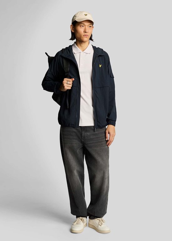 Een man met een pet draagt een lichtpaars poloshirt, een marineblauwe jas, een donkergrijze jeans en een rugzak.