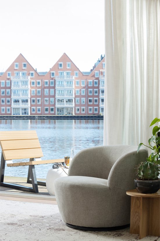 Een lichte fauteuil met een ronde vorm, gestoffeerd in een zandkleurige stof, voor een raam met uitzicht op water en gebouwen.