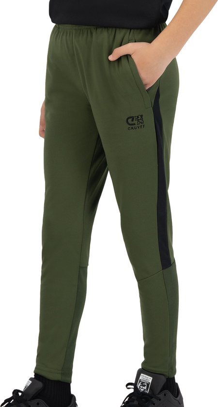 Kind draagt een olijfgroene trainingsbroek met zwarte streep op de zijkant en Cruyff-logo.