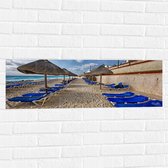 WallClassics - Muursticker - Blauwe Ligbedden op Strand met Rieten Parasols - 90x30 cm Foto op Muursticker