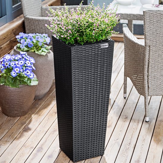 Donkergrijze polyrotan bloempot met gevlochten textuur, gevuld met roze bloemen, op een houten terras.