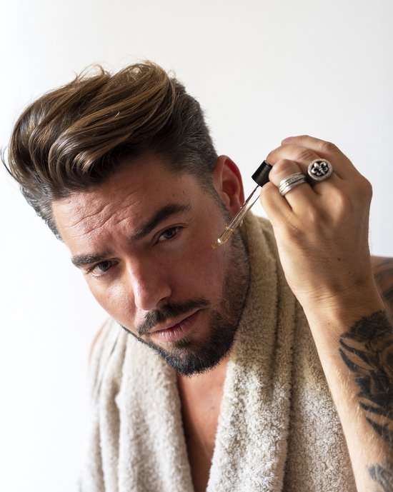 Man met getatoeëerde armen en zilveren ringen die baardolie aanbrengt met een druppelaar.