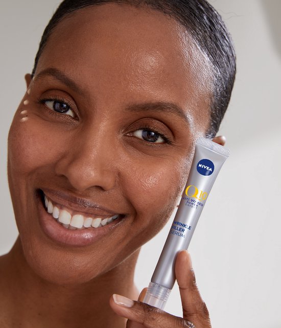 Vrolijke vrouw met donkere huid houdt NIVEA Q10 Anti-Rimpel Expert serum tube vast, met kleine productdruppels op haar gezicht.
