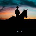 Silhouet van een ruiter op een paard bij zonsondergang, met oplichtende blauwe banden om de benen van het paard.