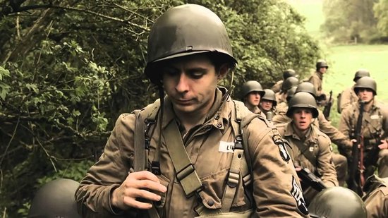 Soldaat in een helm en uniform loopt, met een groep soldaten op de achtergrond in een bosrijke omgeving.