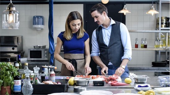Een vrouw in blauw shirt en een man met vest snijden samen aardbeien in een keuken.