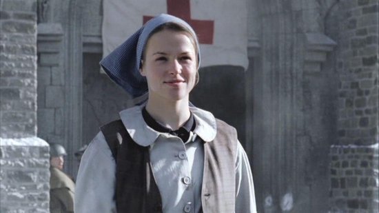 Jonge vrouw in een blauwe hoofddoek en bruin vest lacht, met een rode kruisvlag op de achtergrond.