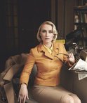 Vrouw met blond haar, zittend aan een bureau in een gele blazer, houdt een bril vast.