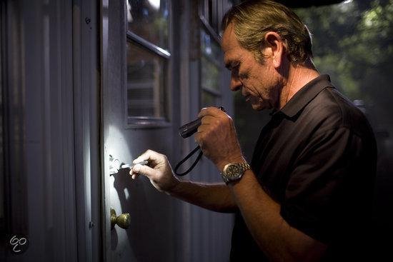 Tommy Lee Jones, met zaklamp in de hand, kijkt naar een slot op een deur.