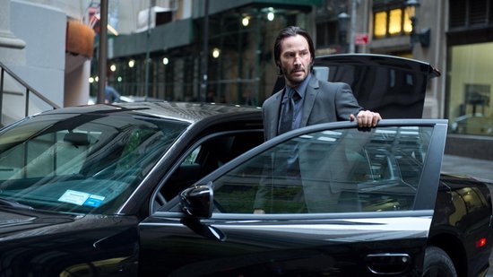 Keanu Reeves in een grijs pak, leunend op een open autodeur.