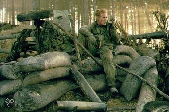 Soldaat zit op zandzakken en praat in een radio, met een gecamoufleerd voertuig en bos op de achtergrond.
