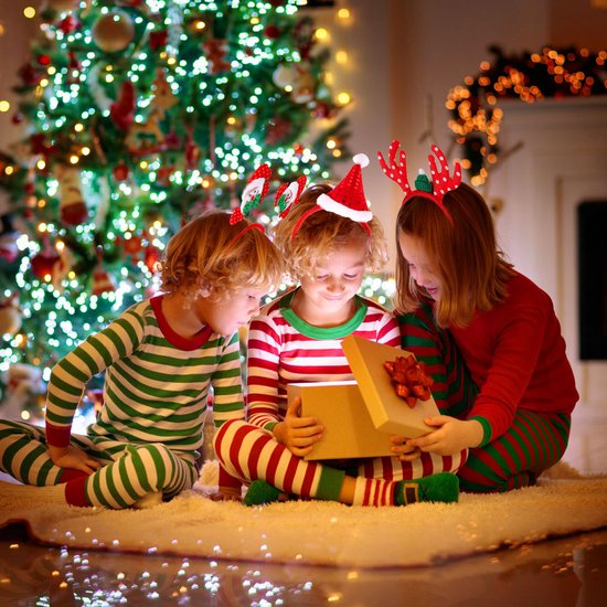 Trois enfants en pyjama ouvrent un cadeau de Noël près du sapin. Tous portent des serre-têtes de Noël.
