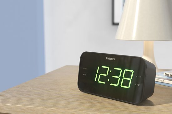 Zwarte Philips wekkerradio met groene digitale tijd op een houten tafel naast een lamp.